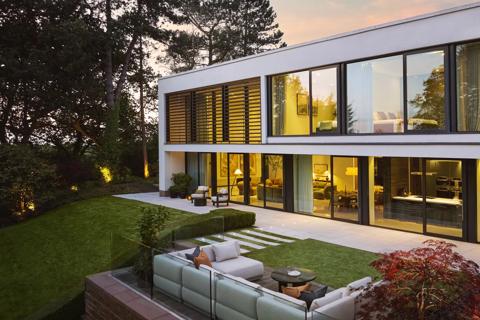 A photo of the house taken from the garden at sunset. The house is lit up inside with warm lighting and you can see into the rooms on both floors through the large windows. In the foreground is the garden furniture.