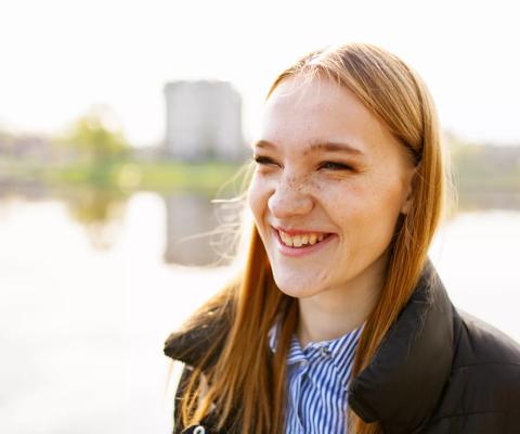 Close up of a young woman's face smiling. She has red hair and freckles, and is standing outside