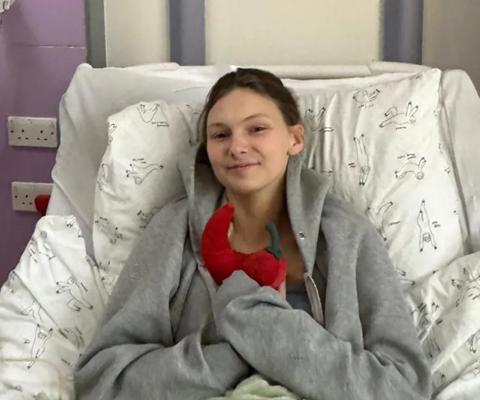 A young woman lying in a hospital bed, smiling and holding a toy red chilli