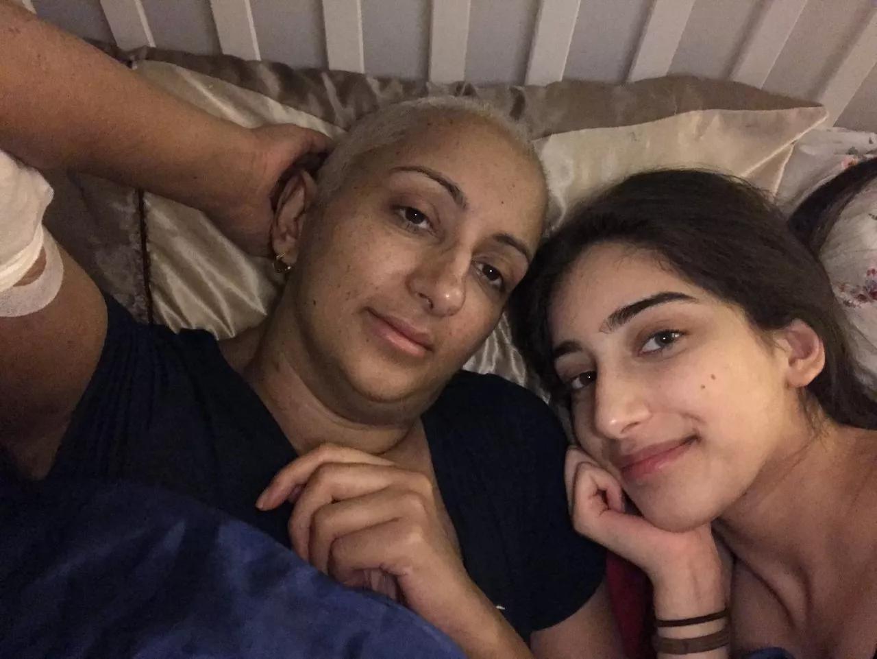 Kal, a South Asian woman with shaved hair, with her teenage daughter, both are lying on a bed smiling