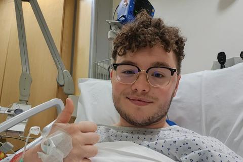 A young man with brown hair and glasses, he's in a hospital bed with a white sheets pulled up to his chest, he's giving a thumbs up