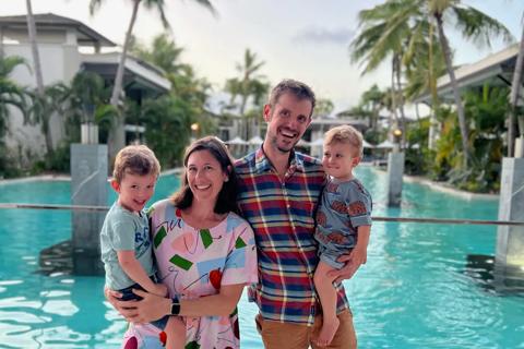 Jake and his wife holding their two sons, standing in front of a pool and palm trees