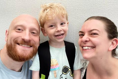 a smiling 3 year old blonde boy stands in the middle of his parents, mum on the right, dad on the left