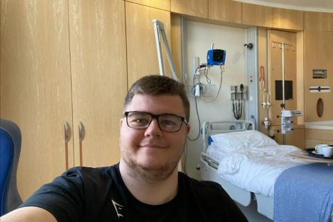 A young man taking a selfie in a hospital room, he has glasses, short brown hair and is wearing a black t-shirt