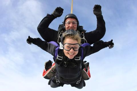 Lady smiling widely, wearing a black parachuting outfit and her arms outstretched on both sides with thumbs up. There is also a skydiver coordinator behind her  with arms up and in a black parachuting outfit too. They are both diving from the sky.