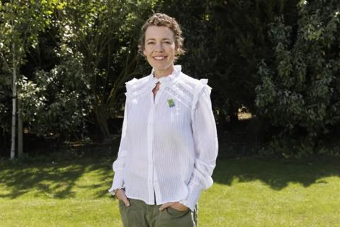 A woman with short brown curly hair and a white blouse is stood in a sunny garden. She is wearing an Anthony Nolan pin.