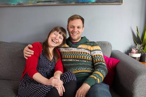 Husband with his arm around his wife sat on their sofa smiling and laughing