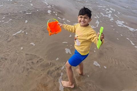 Picture of five-year-old Eesa Hussain on the beach. Eesa is holding a red bucket and green spade as he stands on the shore. 