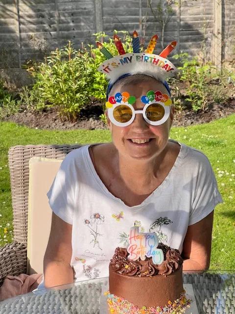 a woman sat in front of a chocolate birthday cake wearing a birthday headband and glasses