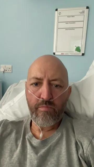 A man with a bald head, grey beard and grey t-shirt with a tube in his nose and white pillow behind him