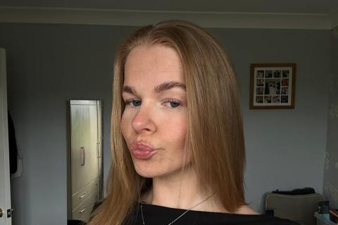 A young white woman is stood in a living room and pouting at the camera.