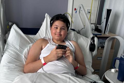 A photo of Reece in a hospital bed, he is holding his phone and wearing black headphones