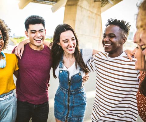 A group of six young people of various different ethnicities laughing and smiling with their arms around each other