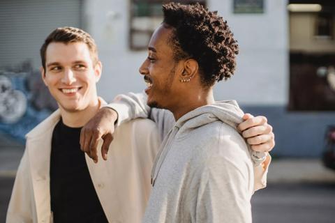 Two guys smiling and as they walk down the street. One of them is a black male wearing a grey hoodie and has an afro hightop. The other is a white male with low cut brown hair, wearing a black top and a cream overshirt