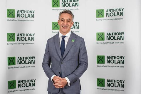 A man in a suit and tie is stood smiling at the camera. He is in front of a backdrop of Anthony Nolan logos.