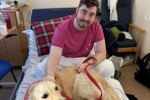 Will, a young man wearing a red t-shirt, sitting in a hospital bed stroking a golden retriever dog