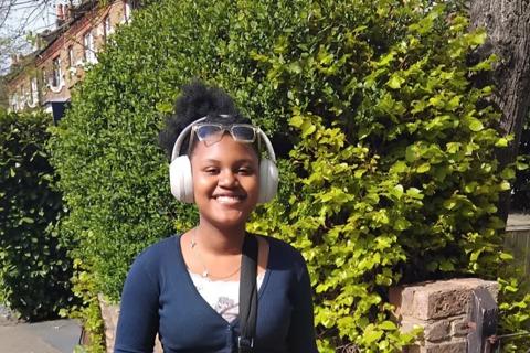 A photograph of Sahara stood outside, she is wearing headphones and a black across-body bag  with different toys and charms attached