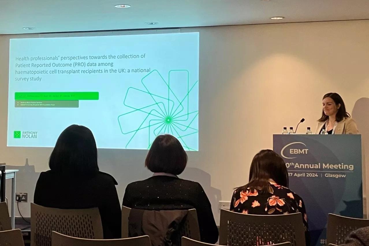 A woman wearing a beige blazer is presenting results from Patient Reported Outcomes research and standing beside a powerpoint being projected on the wall