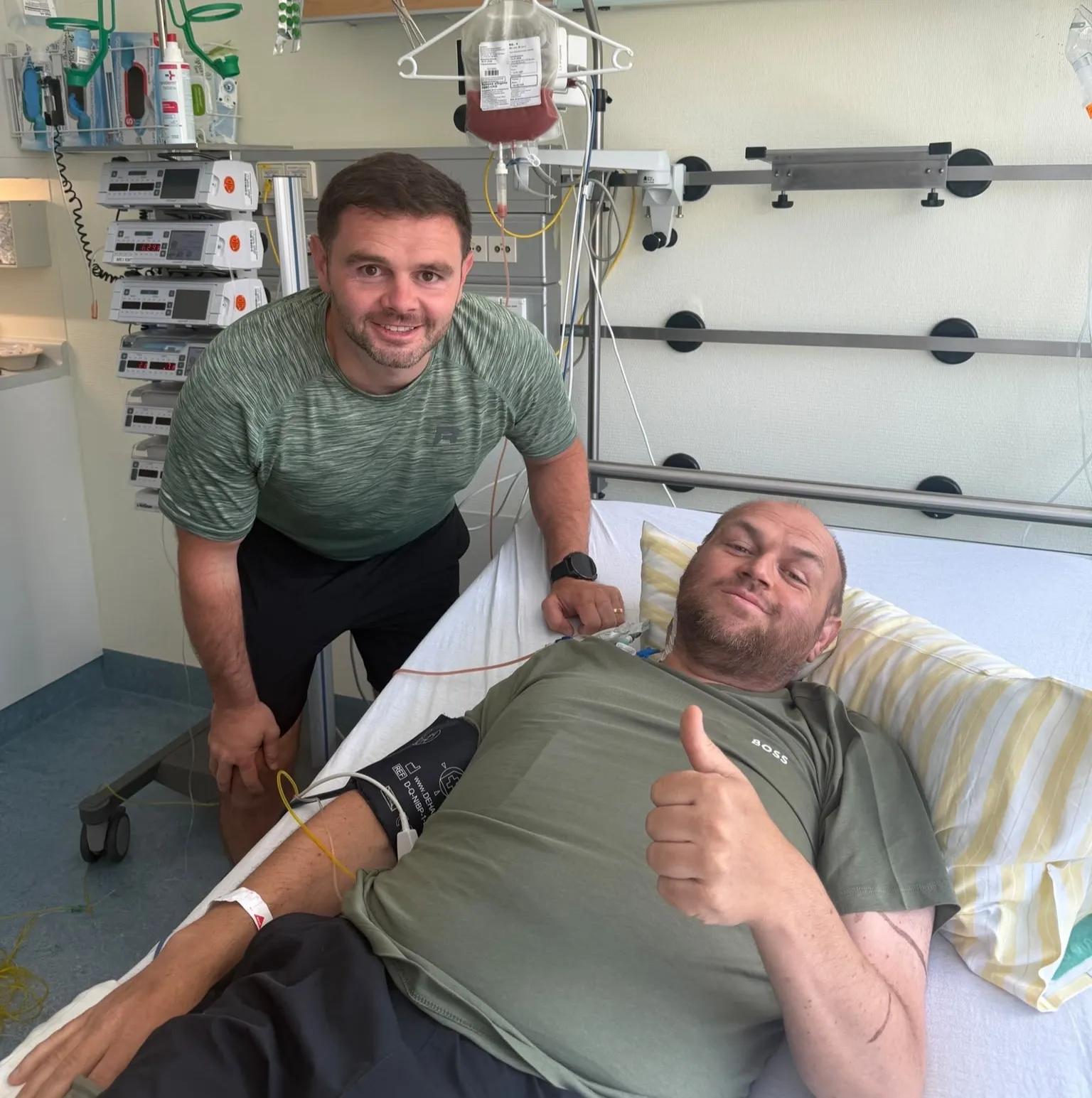Friend smiling stood next to Sean in hospital bed with thumbs up