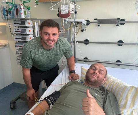 Friend smiling stood next to Sean in hospital bed with thumbs up