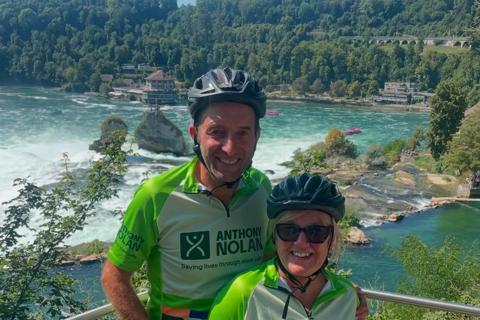 A man and woman wearing Anthony Nolan branded cycling tops and helmets standing in front of a beautiful view of the river Rhine