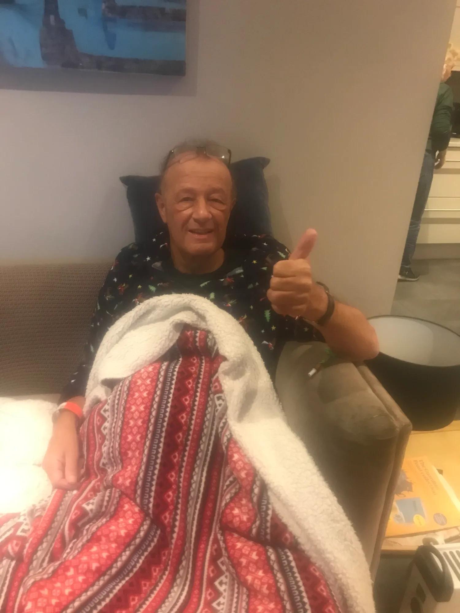 a man sitting on the sofa with a blanket over him giving a thumbs up
