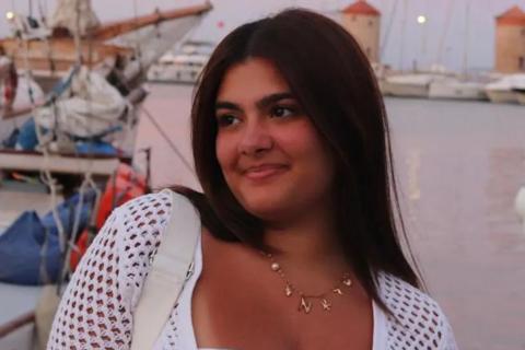 A 19 year old woman with dark hair standing in front of a harbour looking into the distance