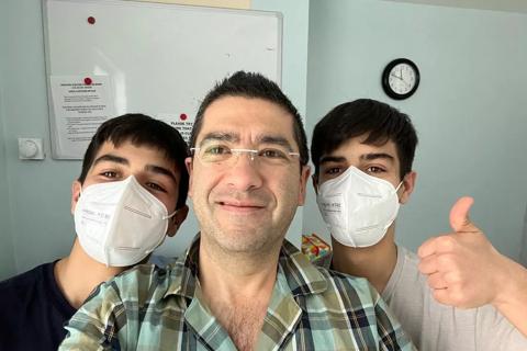 A man with dark hair and glasses and a green checked shirt smiles at the camera, either side of him are his teenage sons also with dark hair and wearing white marks over their nose and mouths, the one on the right is giving a thumbs up