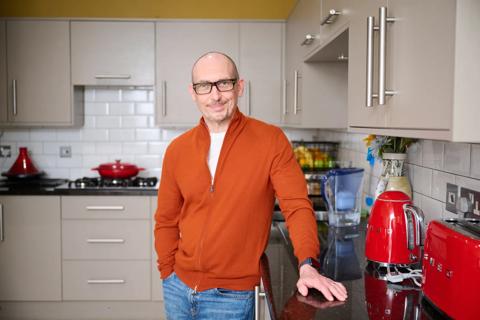 A middle aged white man smiling wearing black thick rim glasses and an orange jumper stood in his kitchen