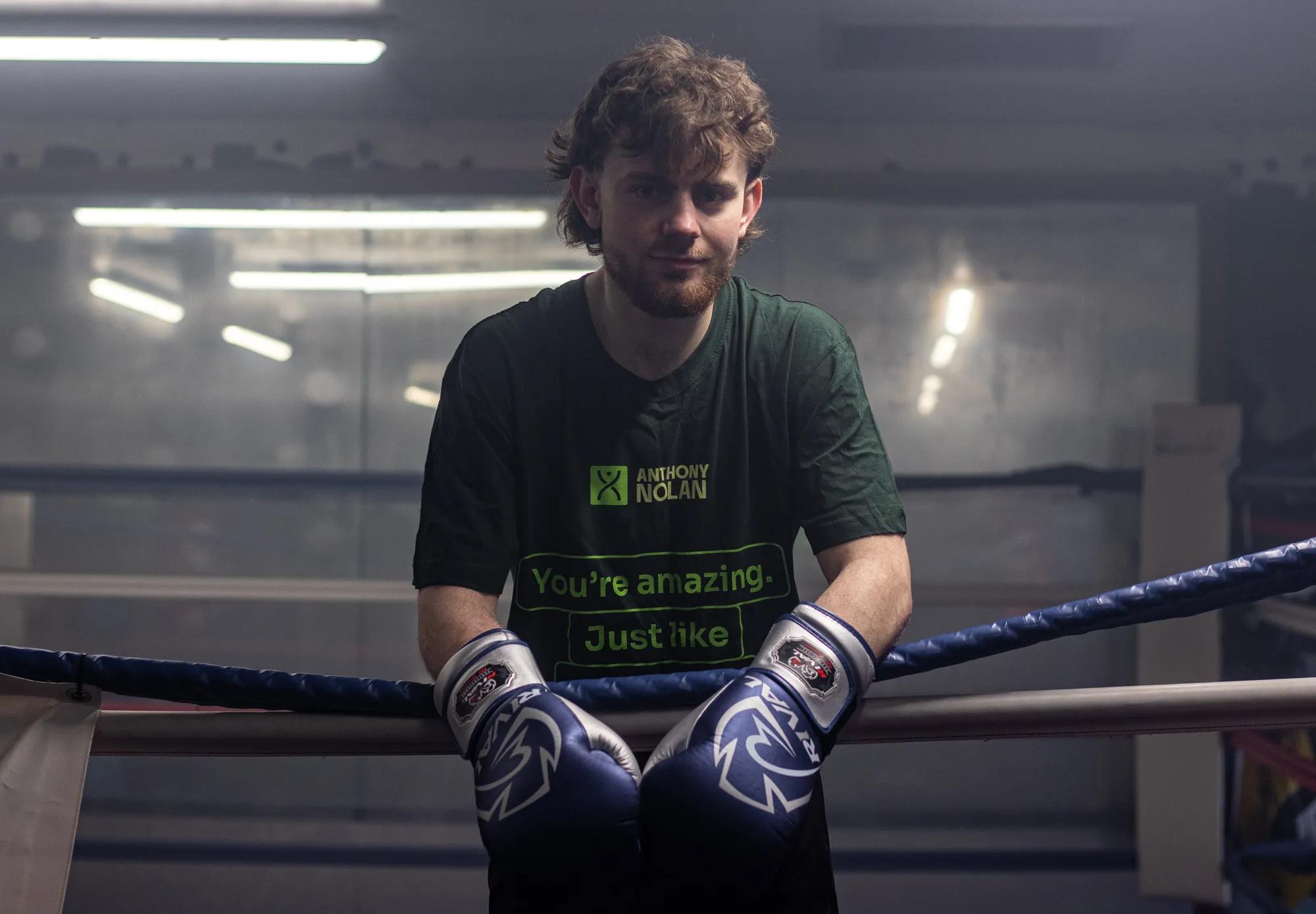 A young man, named Reuben standing in a boxing ring, leaning over the ropes and looking out to camera