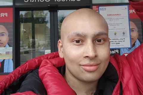 A young man with no hair, wearing a red jacket, he's giving a peace sign outside the cancer centre
